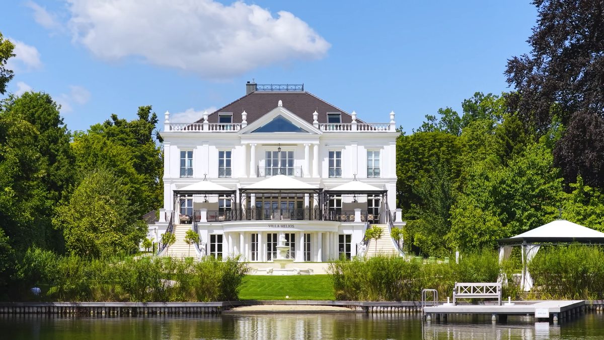 Villa Helios seen from Lake Glindow on a sunny day: white neoclassical façade with terrace pavilions, lawn down to the water